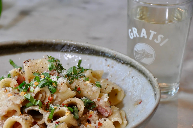 A bowl of rigatoni pasta garnished with chopped herbs, grated cheese, and pepper sits next to a glass of clear water labeled GRATSI on a marble table.