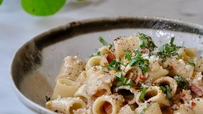 A ceramic bowl filled with rigatoni pasta topped with grated cheese, chopped herbs, pepper, and red pepper flakes, placed on a light-colored table with some greenery in the background.