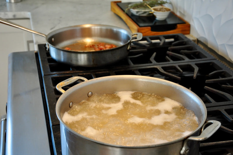 A pot of water boiling on a stove burner in the foreground, with another pan containing food cooking on a back burner; kitchen counter and utensils are visible in the background.