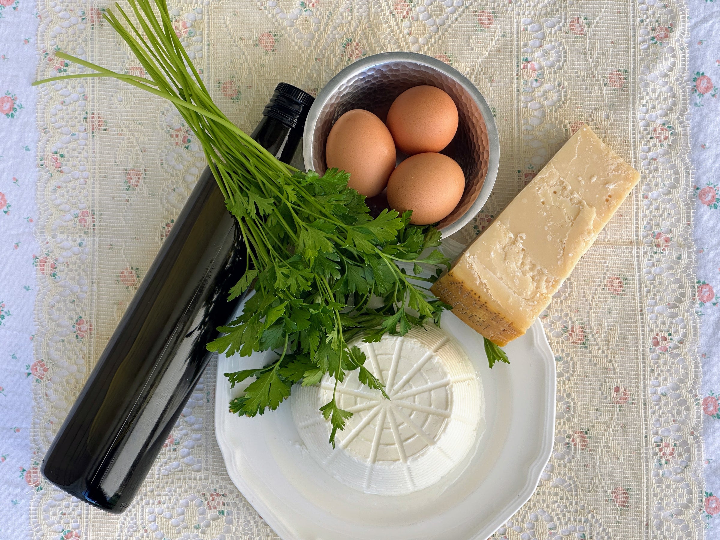 A bottle of olive oil, a bunch of parsley, three brown eggs in a bowl, a wedge of Parmesan cheese, and a wheel of ricotta cheese are arranged on a lacy tablecloth.