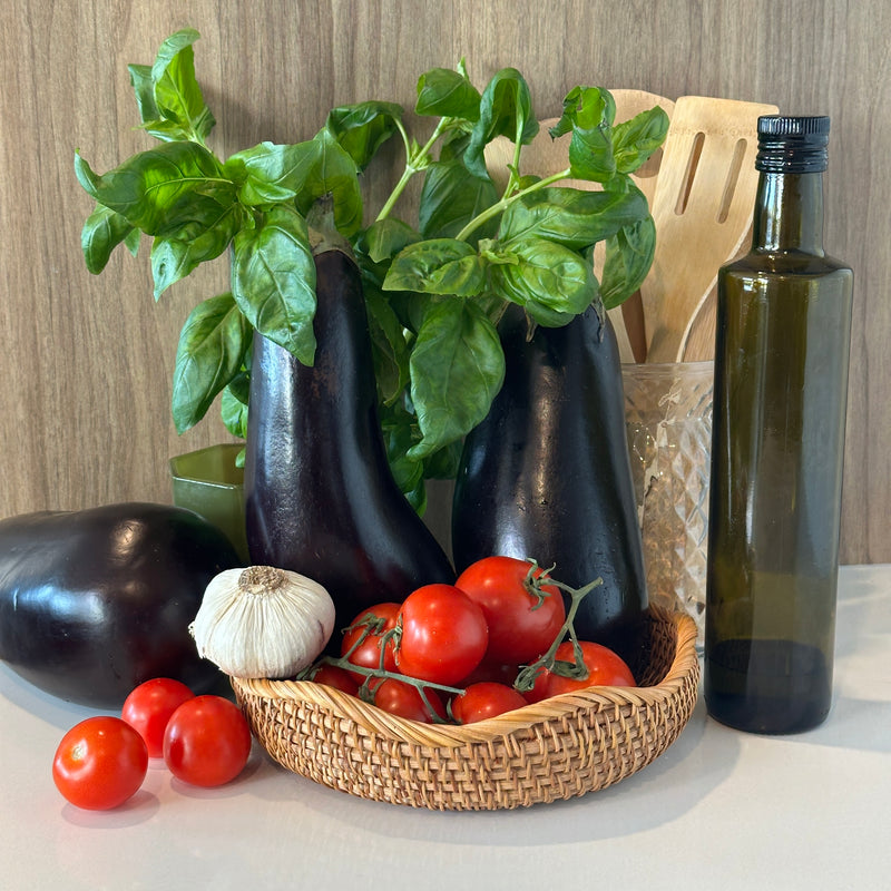 A basket of tomatoes, garlic, and eggplants sits on a counter next to a bottle of olive oil, a basil plant, and wooden utensils in a glass container, with a wooden background.
