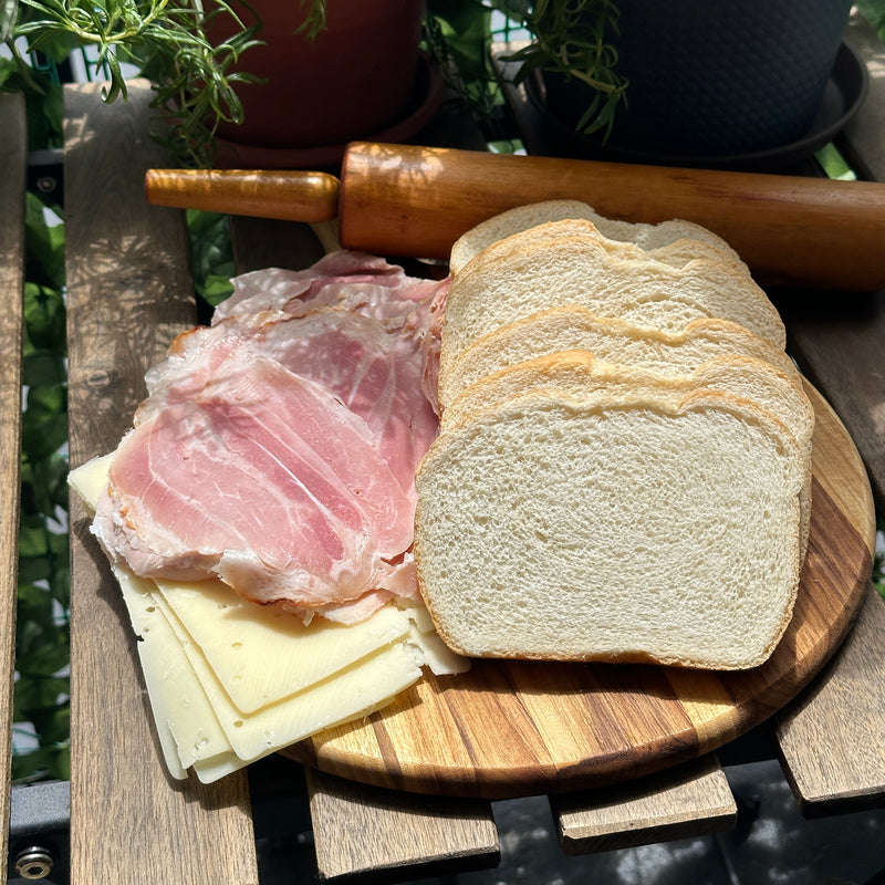 Slices of white bread, ham, and cheese are arranged on a round wooden board. A rolling pin sits behind them, all placed on a wooden surface with potted plants nearby and sunlight streaming in.