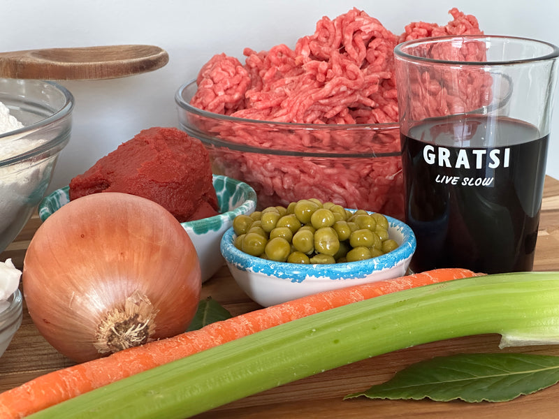 A close-up of raw ground beef, a glass of red wine, an onion, a carrot, a celery stalk, tomato paste, green peas, and a bay leaf arranged on a wooden surface. The wine glass says GRATSI LIVE SLOW.
