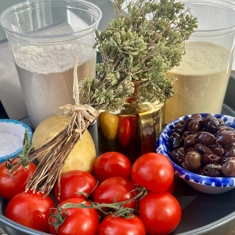 A tray with fresh tomatoes, a potato, black olives in a blue bowl, a bunch of dried herbs, a metal container, and two plastic cups filled with flour and cornmeal.