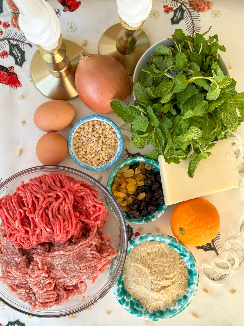 A top view of ingredients on a table, including ground meat, eggs, an onion, parsley, mint, cheese, breadcrumbs, chopped nuts, raisins, and an orange. Two candles and a festive tablecloth are also visible.