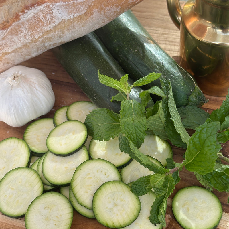 A wooden board with sliced zucchini, whole zucchinis, a bunch of fresh mint, a garlic bulb, a loaf of crusty bread, and a bottle of olive oil.