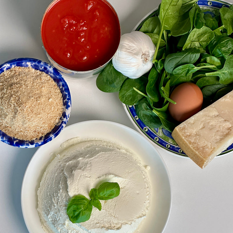 A top-down view of ricotta cheese with basil, canned tomatoes, fresh spinach, a garlic bulb, an egg, Parmesan cheese, and breadcrumbs arranged on a white surface.
