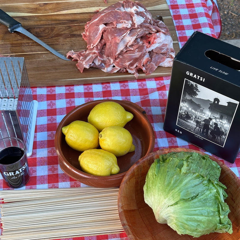 A table with sliced raw meat, a box of Gratsi red wine, a glass of wine, fresh lemons in a bowl, uncooked pasta, and a head of lettuce on a wooden bowl atop a red checkered tablecloth.