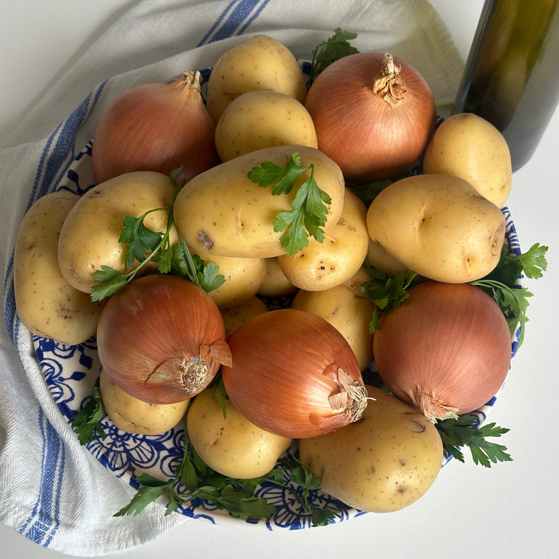 A blue and white patterned bowl filled with whole yellow potatoes, brown onions, and sprigs of parsley. A white cloth with blue stripes and a green glass bottle are beside the bowl on a white surface.
