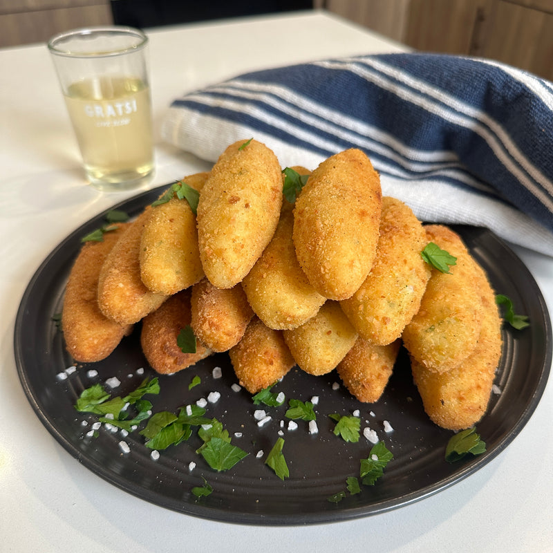 A black plate stacked with golden, breaded croquettes garnished with chopped parsley and coarse salt. A glass of white wine and a blue-striped cloth are in the background on a white countertop.