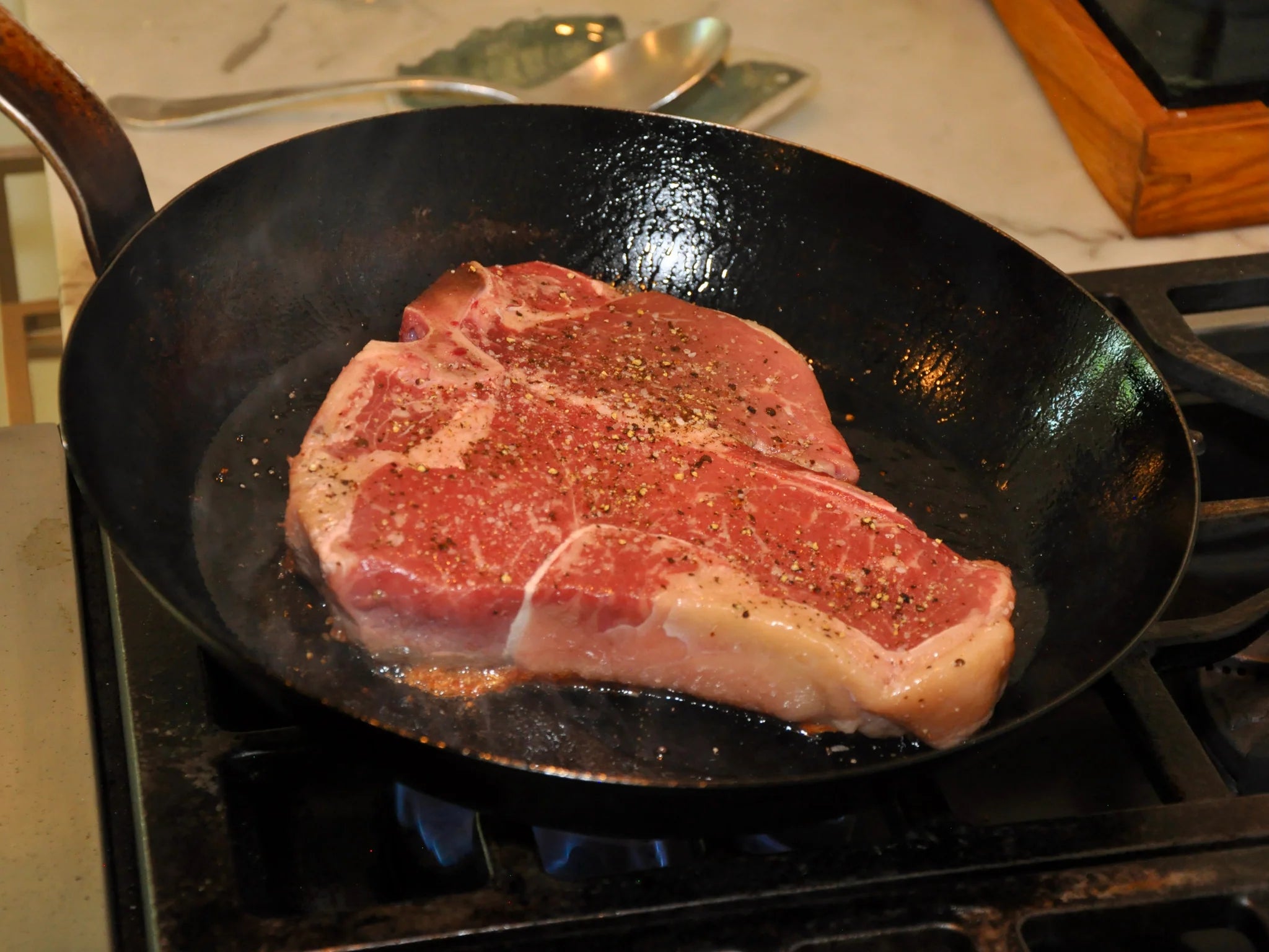 A raw, seasoned T-bone steak sizzles in a black cast iron skillet on a stove, ready to be cooked. Steam rises from the pan, and a spoon rests on the countertop in the background.