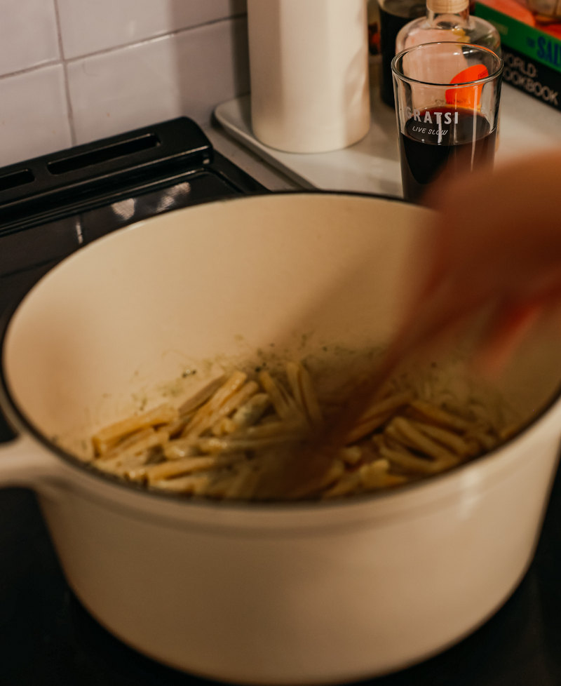 A hand stirs pasta in a cream-colored pot on a stove. In the background, there is a glass of dark beverage and some kitchen items.