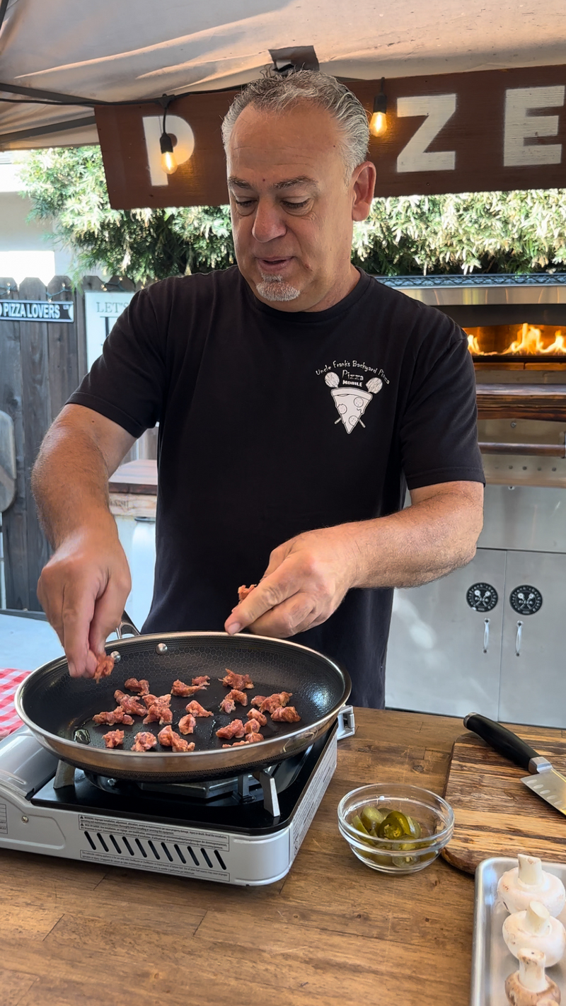 A man cooks sliced meat in a frying pan on a portable stove outdoors, with a wood-fired pizza oven burning in the background. He wears a black T-shirt and stands at a wooden counter with ingredients nearby.