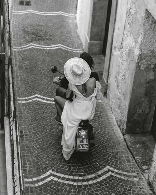 A woman in a white dress and wide-brimmed hat rides on the back of a scooter with another person along a narrow, cobblestone street between old buildings. The photo is in black and white.
