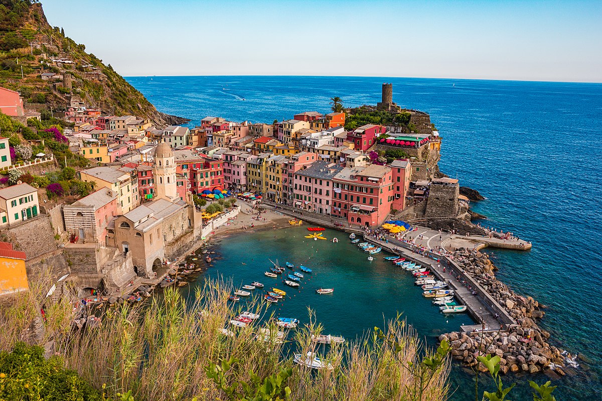Colorful seaside village with clustered buildings on a rocky coast, boats docked in a small harbor, and clear blue sea in the background, viewed from a hillside with greenery in the foreground.