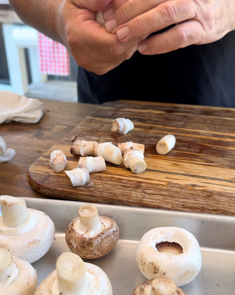 A person’s hands prepare mushrooms on a wooden cutting board, with whole and cut mushrooms visible. The scene suggests food preparation in a kitchen setting.