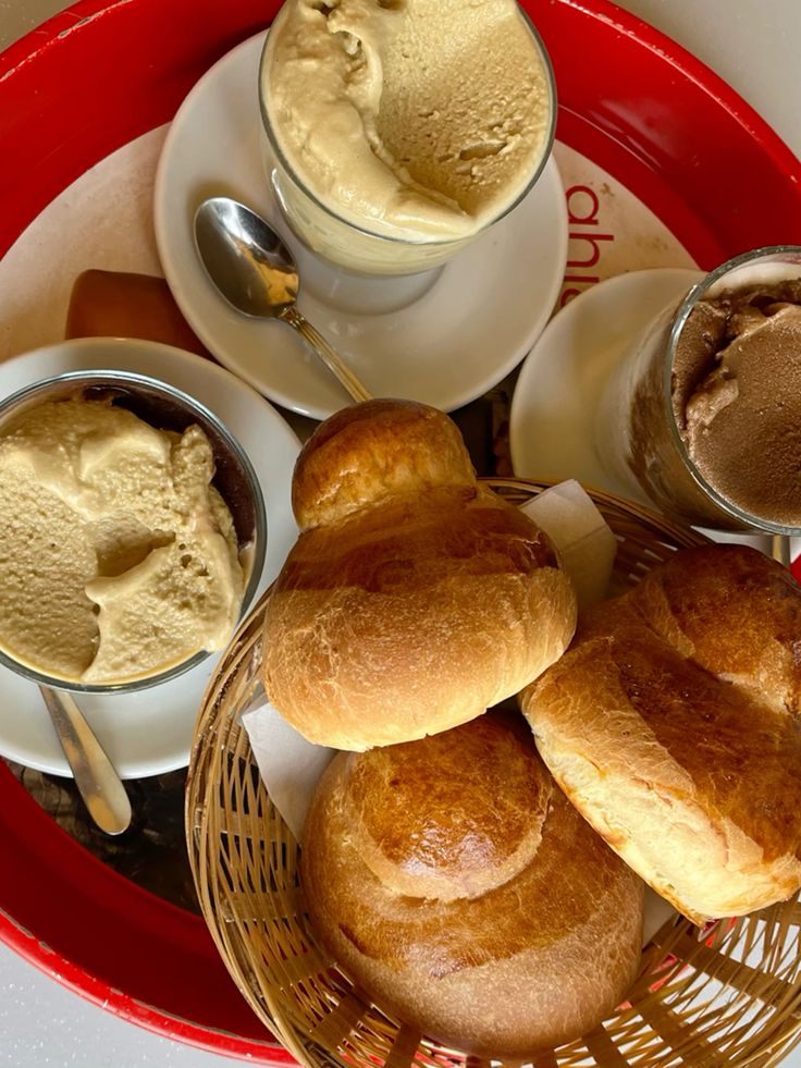 A basket of shiny brioche rolls sits on a red tray beside three glass cups filled with creamy gelato, each on a saucer with a spoon. The setup suggests a classic Italian breakfast or dessert.