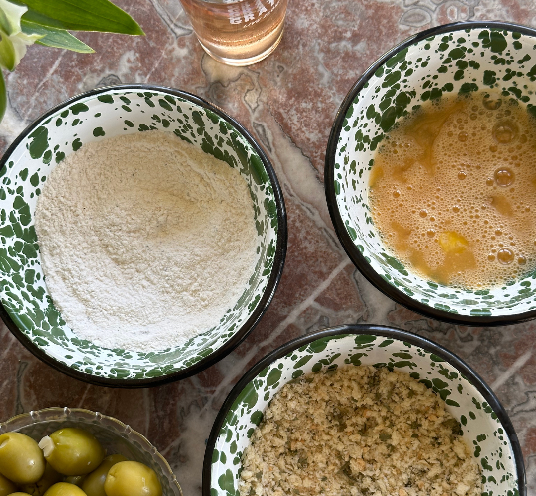 Three bowls on a marbled surface: one with flour, one with beaten eggs, and one with seasoned breadcrumbs. Nearby are green olives in a dish and a glass of rosé wine.