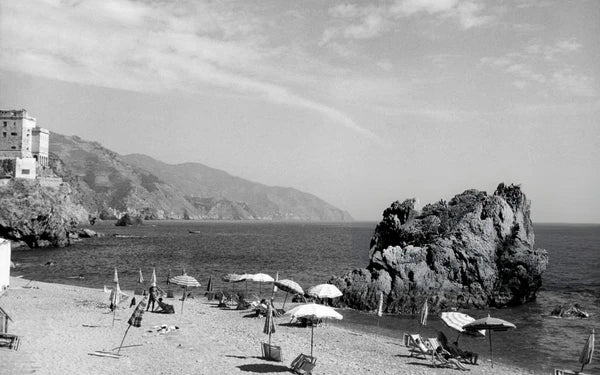 A black-and-white photo of a beach with scattered umbrellas and lounge chairs. Rocky cliffs and mountains line the coast. Calm sea water meets the shore under a partly cloudy sky. Few people are visible, creating a tranquil scene.