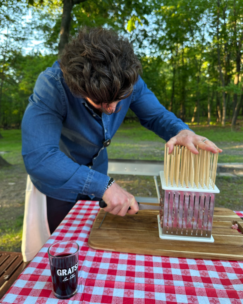 A person in a blue shirt uses a cutting tool with wooden skewers to slice potatoes on a wooden board, outdoors on a picnic table with a red and white checkered tablecloth. A glass with GRATIS text is nearby. Trees are in the background.
