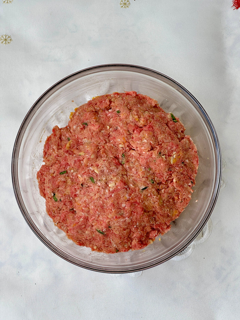 A glass bowl filled with a mixture of raw ground meat, herbs, and seasonings sits on a white surface. The mixture appears ready to be shaped for recipes like meatballs or meatloaf.