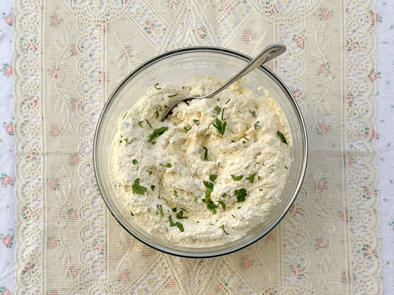 A clear glass bowl filled with a creamy, white mixture garnished with chopped green herbs, sits on a lace-patterned tablecloth. A metal spoon rests inside the bowl.