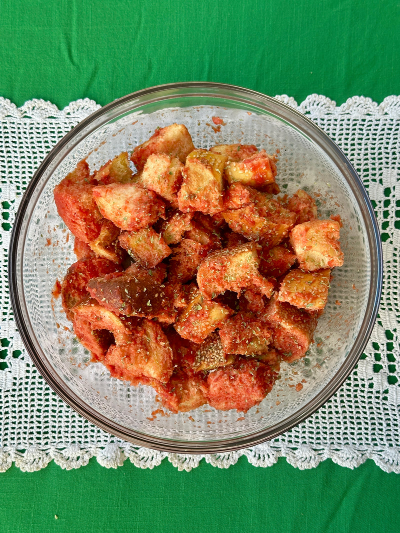 A clear glass bowl filled with seasoned, toasted bread cubes sits on a white lace placemat over a green tablecloth. The bread is coated with red seasoning and herbs.