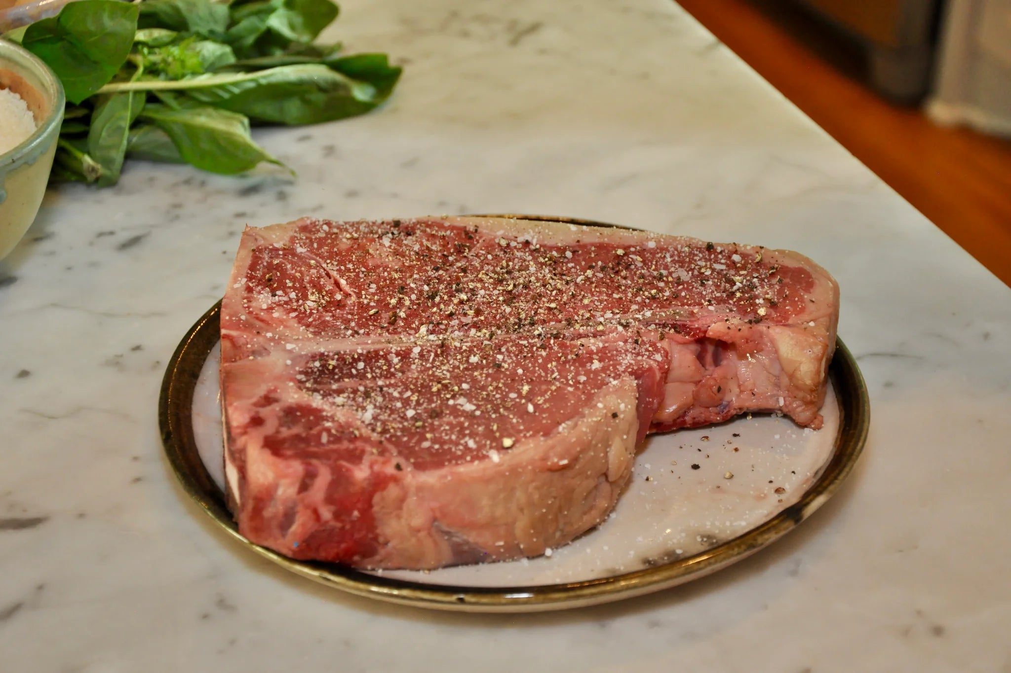 A raw, seasoned T-bone steak sits on a round plate atop a marble countertop, with fresh spinach leaves visible in the background.