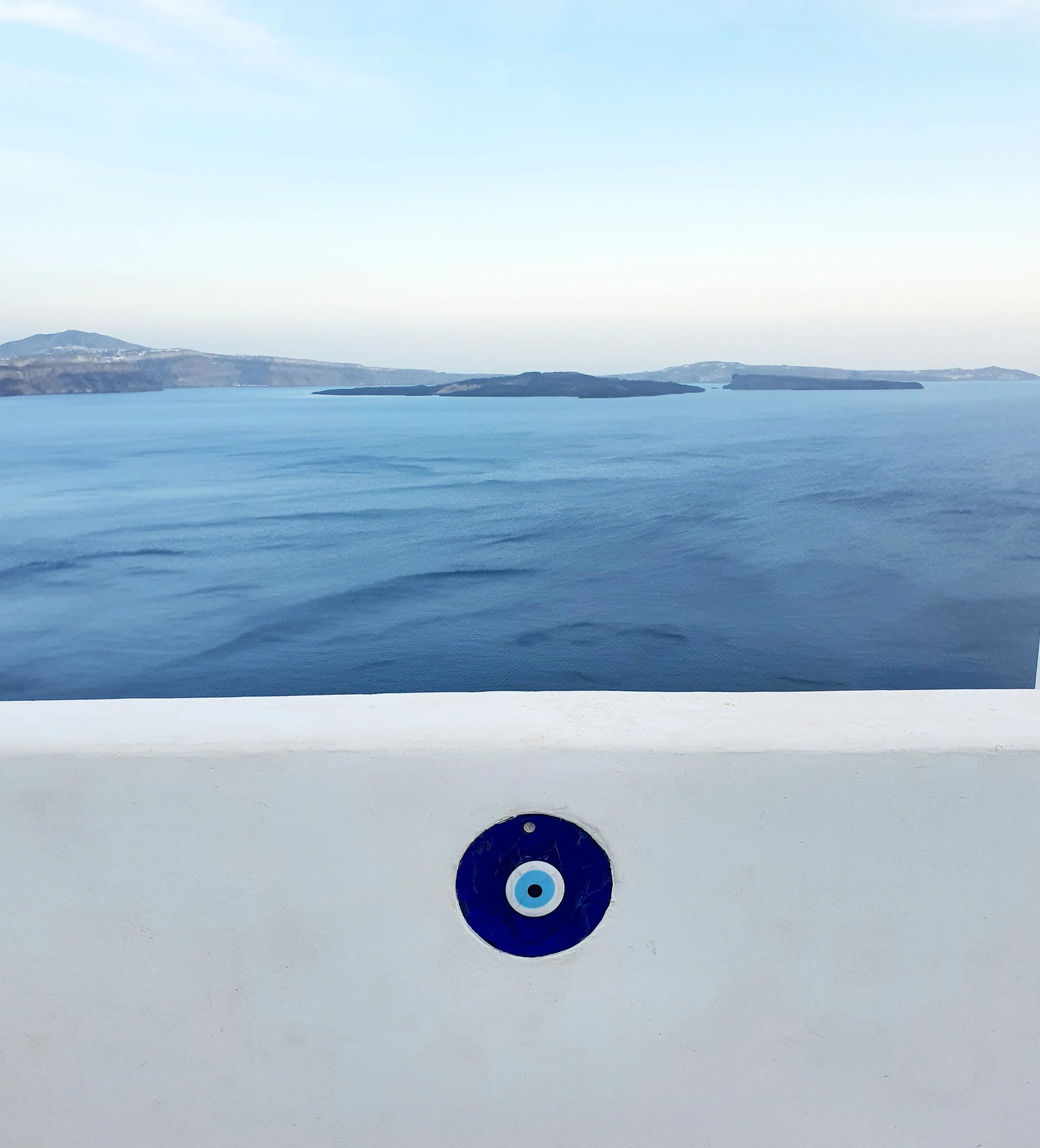A white wall with a blue evil eye charm overlooks a calm blue sea and distant islands under a clear sky.