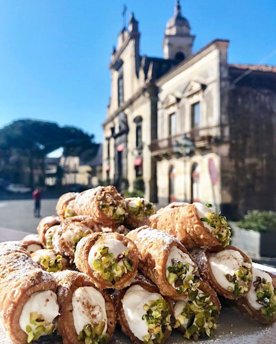A pile of cannoli filled with creamy ricotta and garnished with chopped pistachios sits on a plate outdoors, with an old stone building and blue sky in the background.