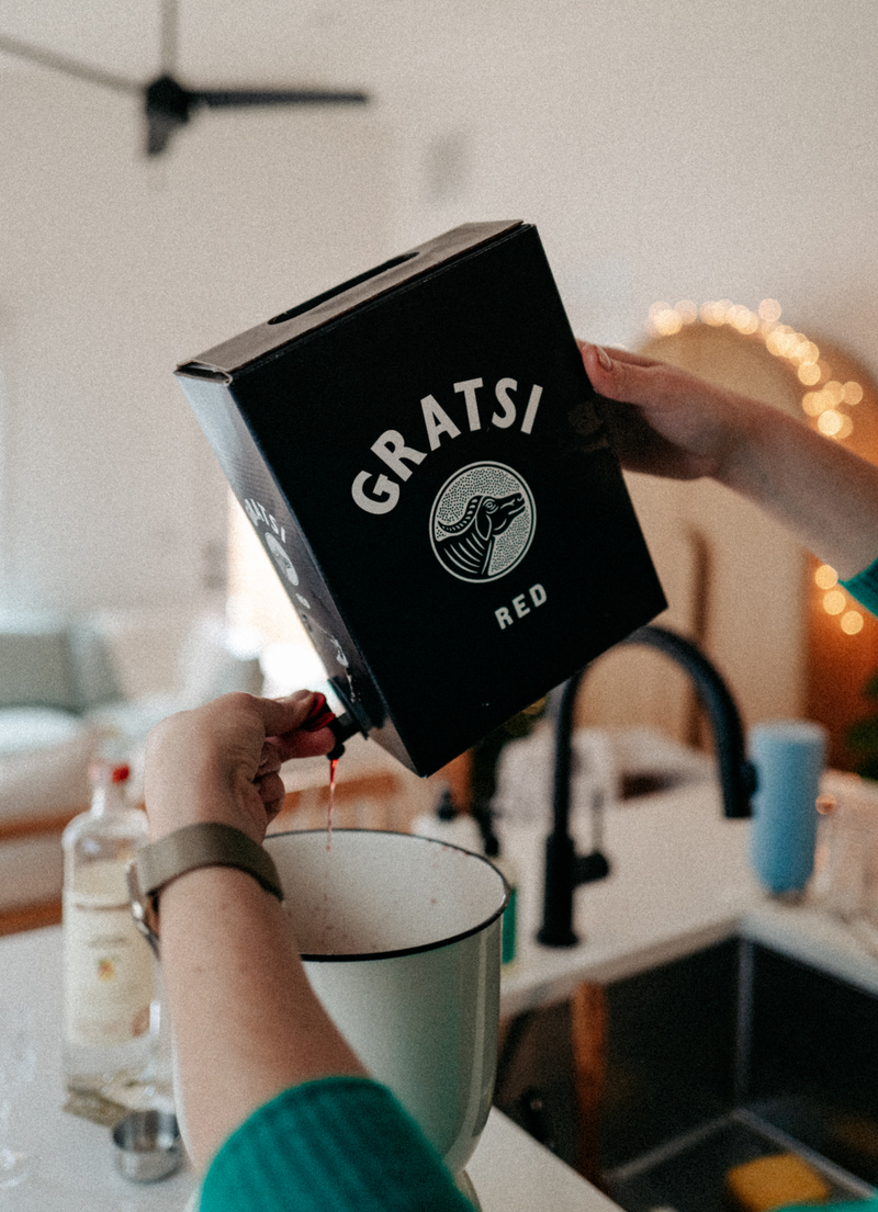 A person pours red wine from a Gratsi Red box into a white pot in a kitchen. The background is softly lit with warm lights and some kitchen items are visible on the counter.