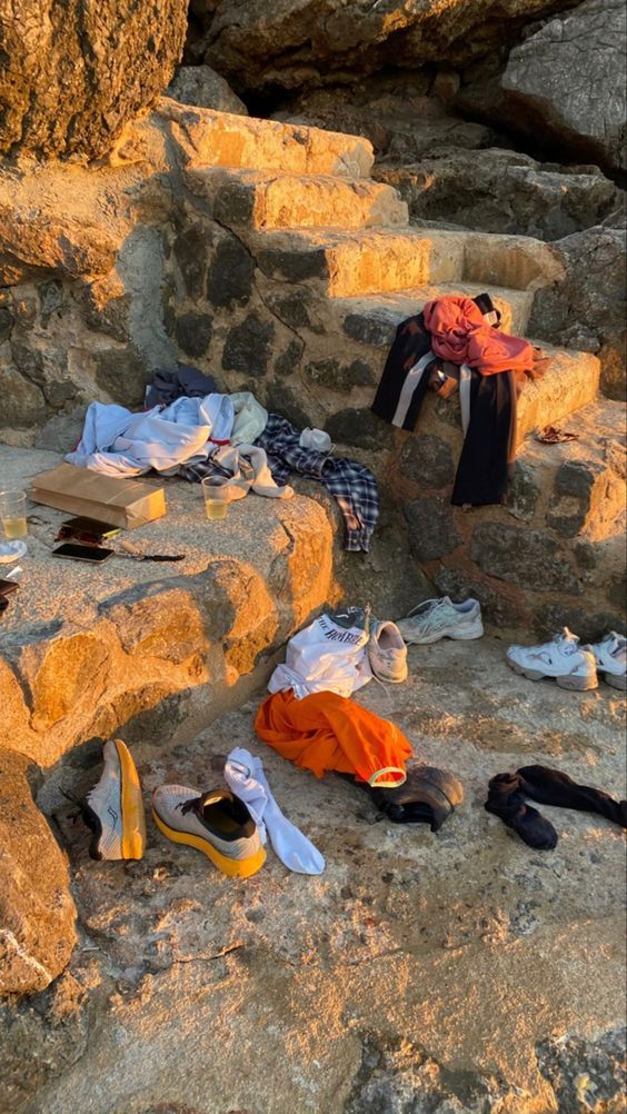 Clothes, shoes, socks, and plastic cups are scattered on stone steps and rocks in an outdoor setting, lit by warm sunlight. The scene suggests a casual, possibly abandoned gathering spot.