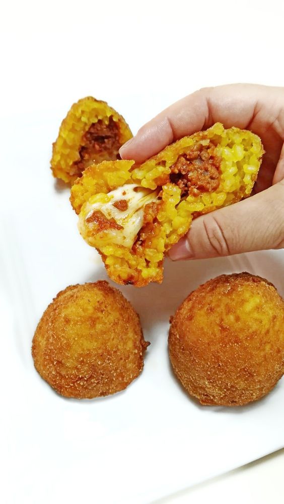 A hand holds a halved arancini, showing a filling of meat sauce and melted cheese. Two whole, golden-brown, breaded rice balls are on a white plate below.