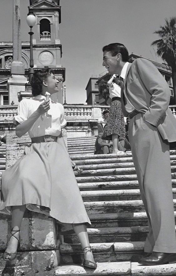 A woman in a skirt and blouse sits on stone steps eating ice cream, smiling at a man in a suit who is standing and leaning toward her. They appear to be chatting happily in a vintage city setting.
