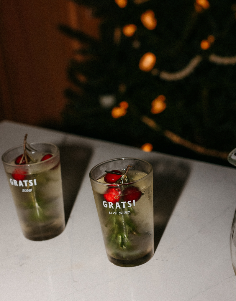 Two glasses with clear drinks, ice, and garnishes of cranberries and rosemary sit on a white surface. The glasses are labeled GRATSI LIVE SLOW. A blurred, decorated Christmas tree is in the background.