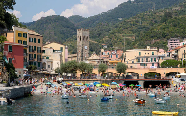 Busy beach scene in an Italian coastal town with colorful umbrellas, people swimming and sunbathing, small boats on the water, vibrant buildings, and green hills with a stone clock tower in the background.