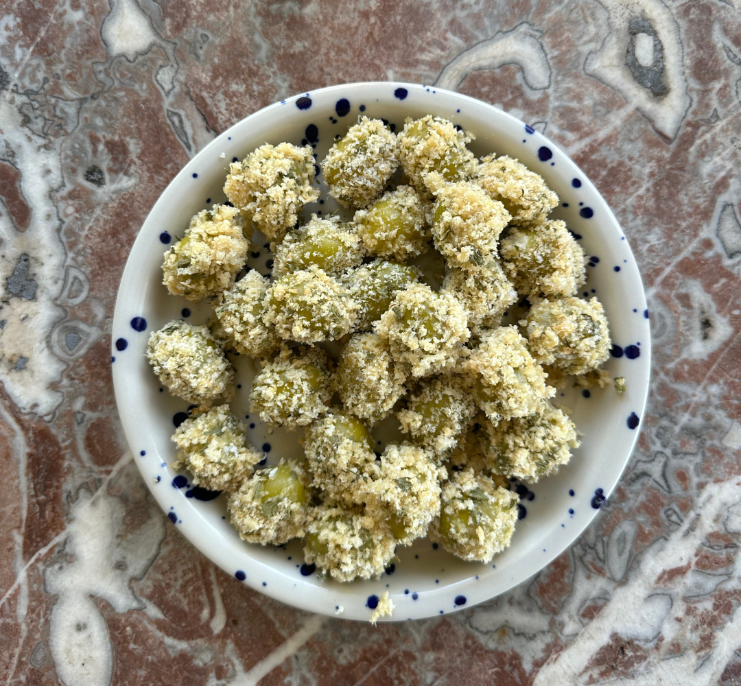 A small white bowl with blue dots holds green olives coated in a crunchy, crumbly breading, sitting on a marbled pink and gray surface.