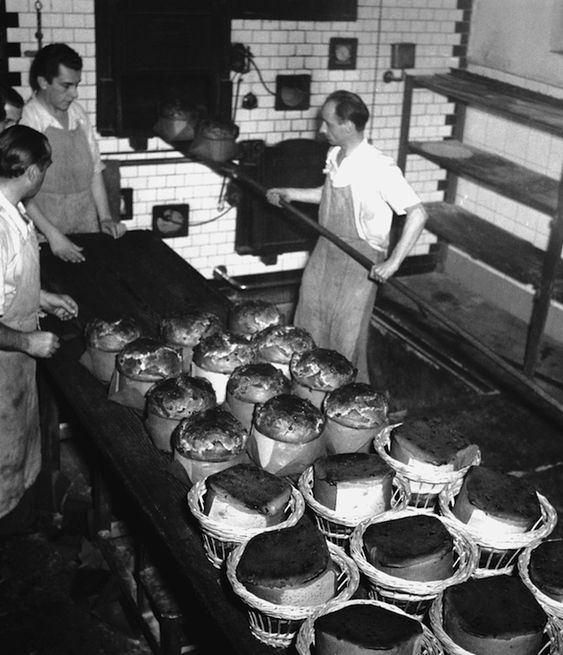 Three men in aprons stand in a bakery, with dozens of large loaves of bread cooling on a long table. The background shows tiled walls, an oven, and empty shelves.