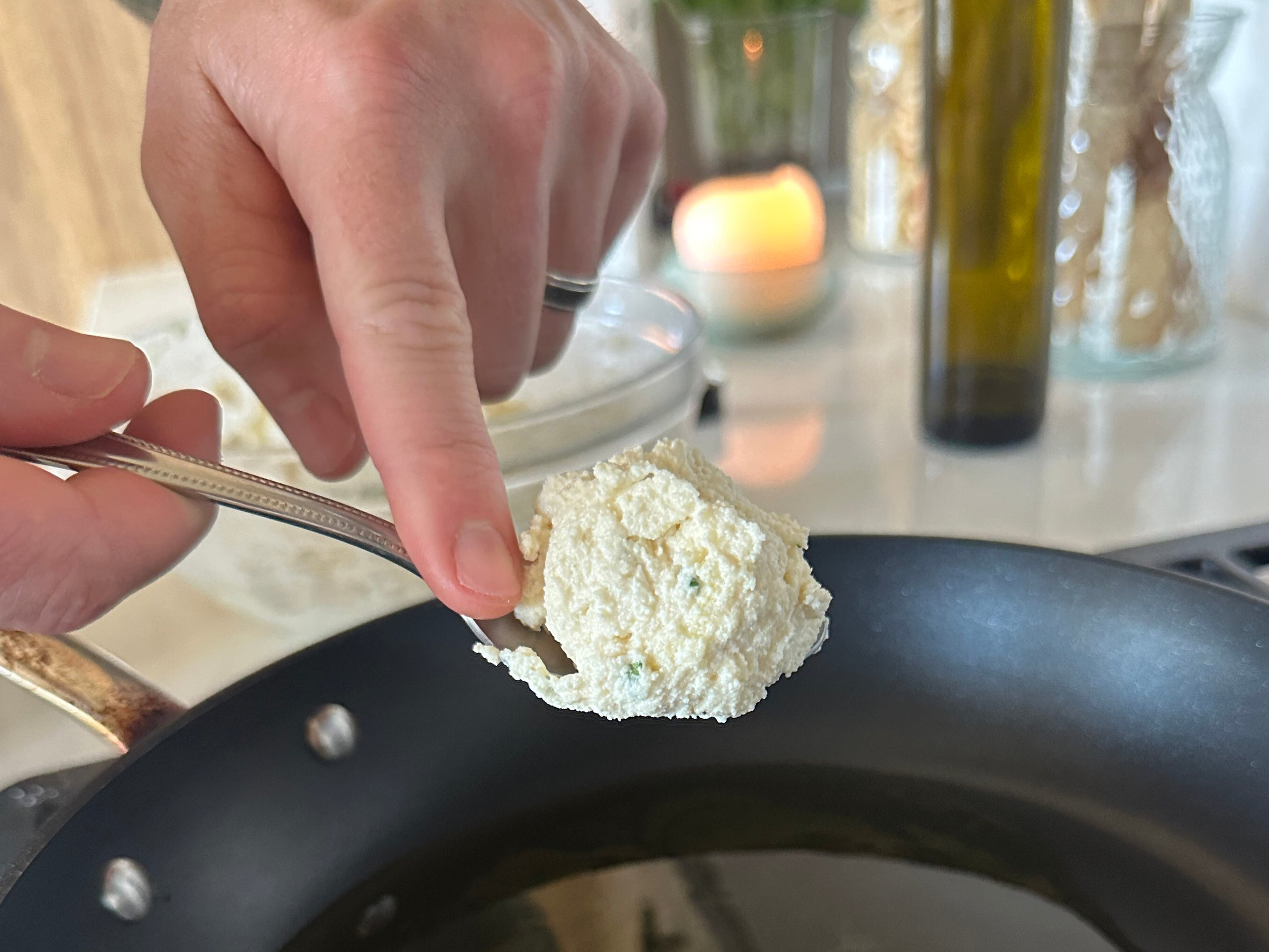 A hand holding a spoonful of ricotta cheese over a skillet on a stove, preparing to cook. Kitchen items and a lit candle are visible in the blurred background.