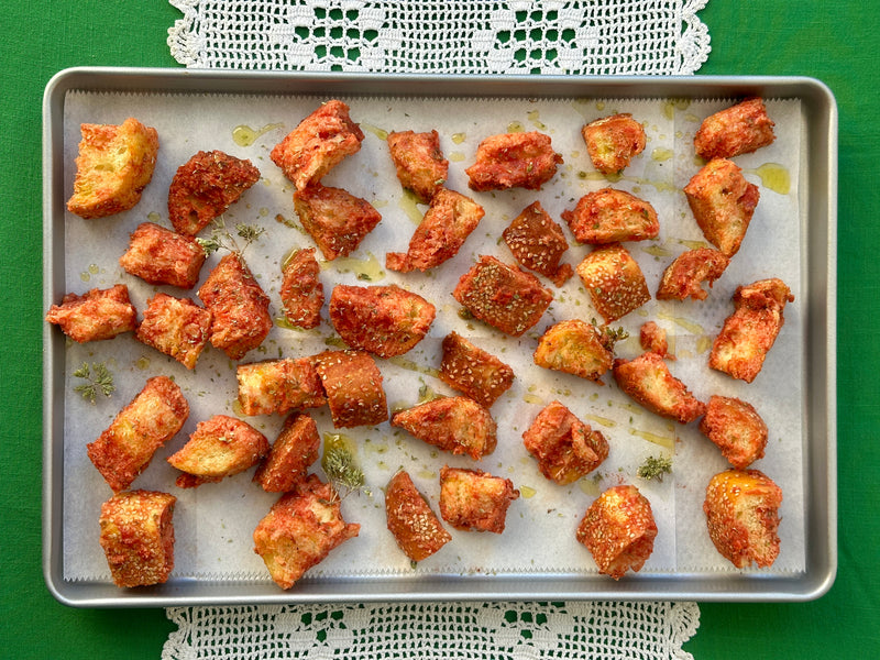 A baking tray lined with parchment paper holds golden brown, seasoned bread cubes spread out evenly. The tray sits on a green tablecloth with a white lace doily underneath.