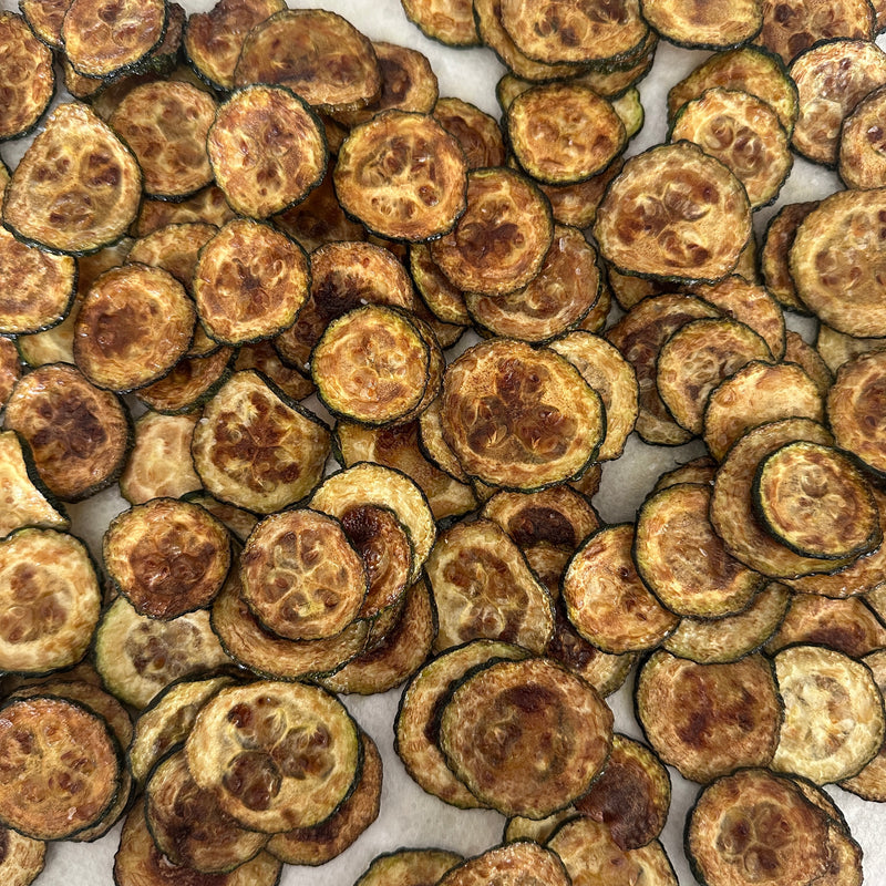 A close-up of many baked or dehydrated zucchini chips arranged in an overlapping pattern, showing golden brown and crispy textures.
