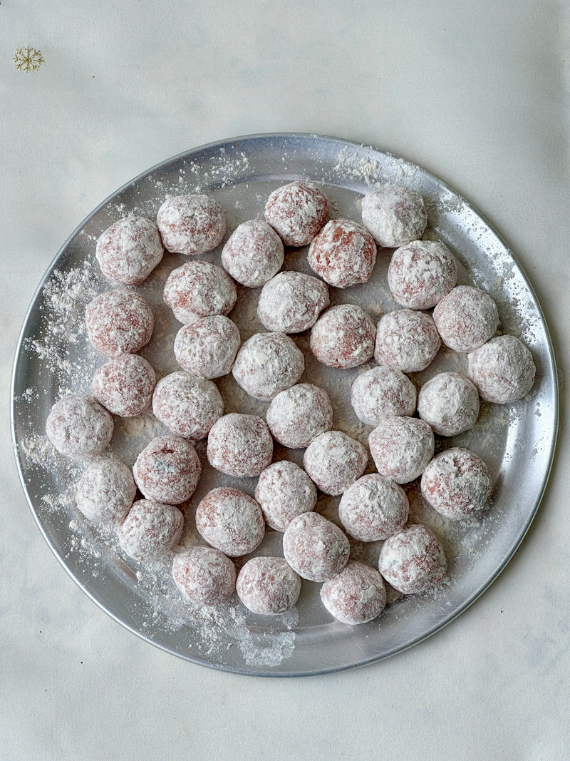 A round metal tray filled with several powdered sugar-coated balls, likely homemade sweets or cookies, arranged on a white surface. Some powdered sugar is scattered on the tray and surface.