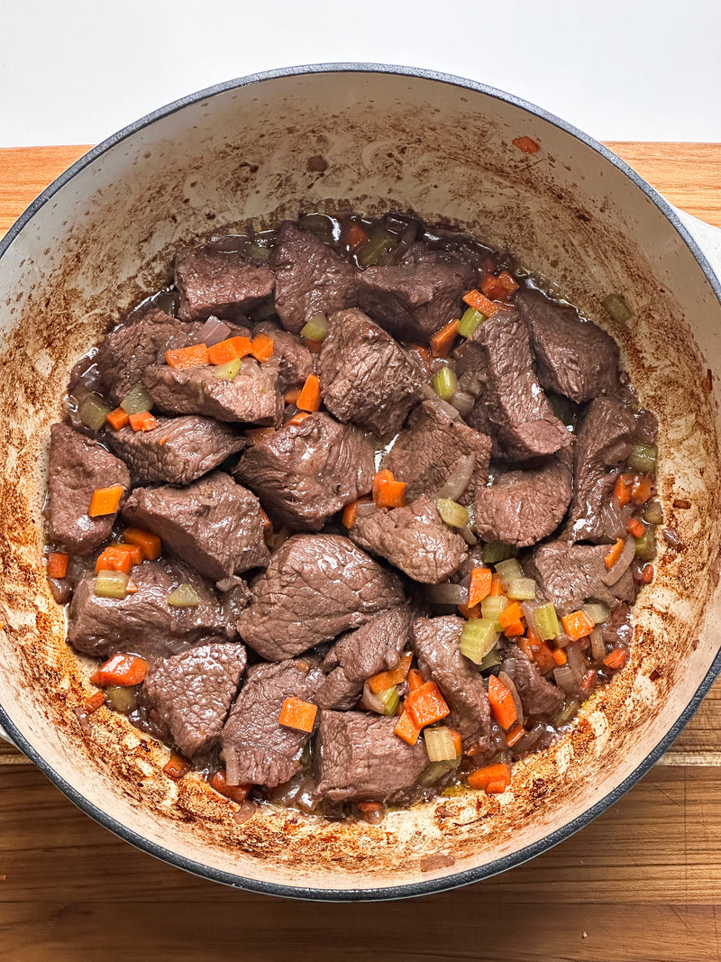 Chunks of browned beef with diced carrots and celery cook in a large, round, enameled pot. The edges of the pot show browned bits, indicating simmering over time. The food appears to be the beginning of a stew.