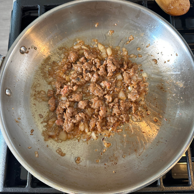 Ground meat and chopped onions are being sautéed in a stainless steel skillet on a stovetop, with some oil and browned bits visible in the pan.