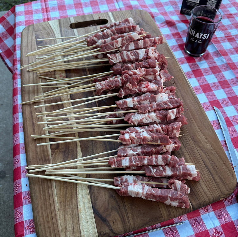 Skewers of raw, cubed meat are arranged on a wooden cutting board atop a red and white checkered tablecloth. A glass of dark liquid and a knife are visible nearby.