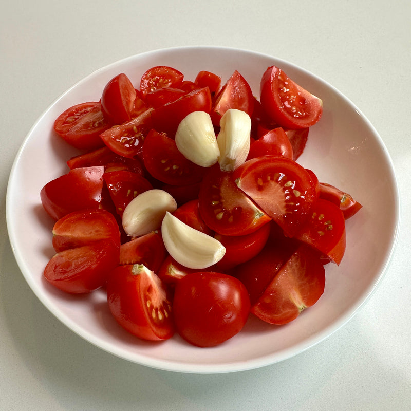 A white bowl filled with quartered red tomatoes and several peeled garlic cloves, placed on a white surface.