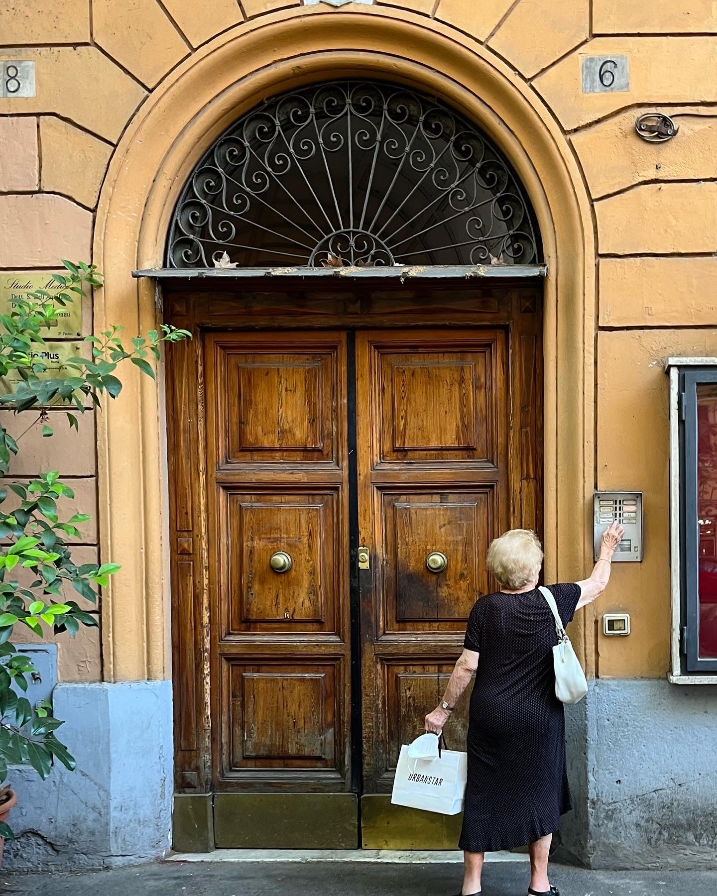 An elderly woman in a black dress stands at a large wooden door, pressing a button on an intercom. She holds a white tote and a shopping bag, and the building has an arched doorway with number 6 above it.