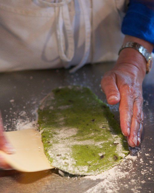A person uses a dough scraper to shape a green dough with dark specks on a floured surface. Only their hands and part of their blue sleeve and white apron are visible.