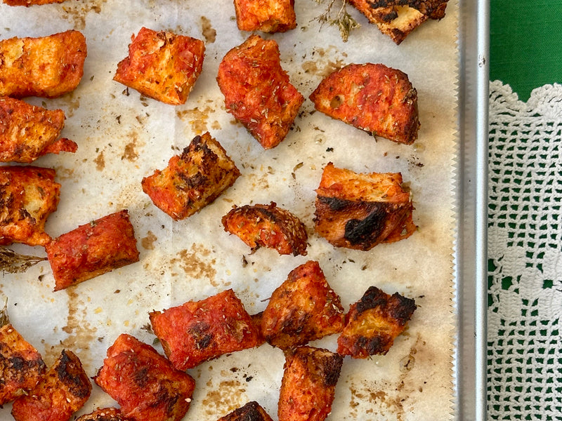 Chunks of roasted, marinated paneer with browned edges on parchment paper atop a baking tray, next to a green tablecloth and a white lace doily.