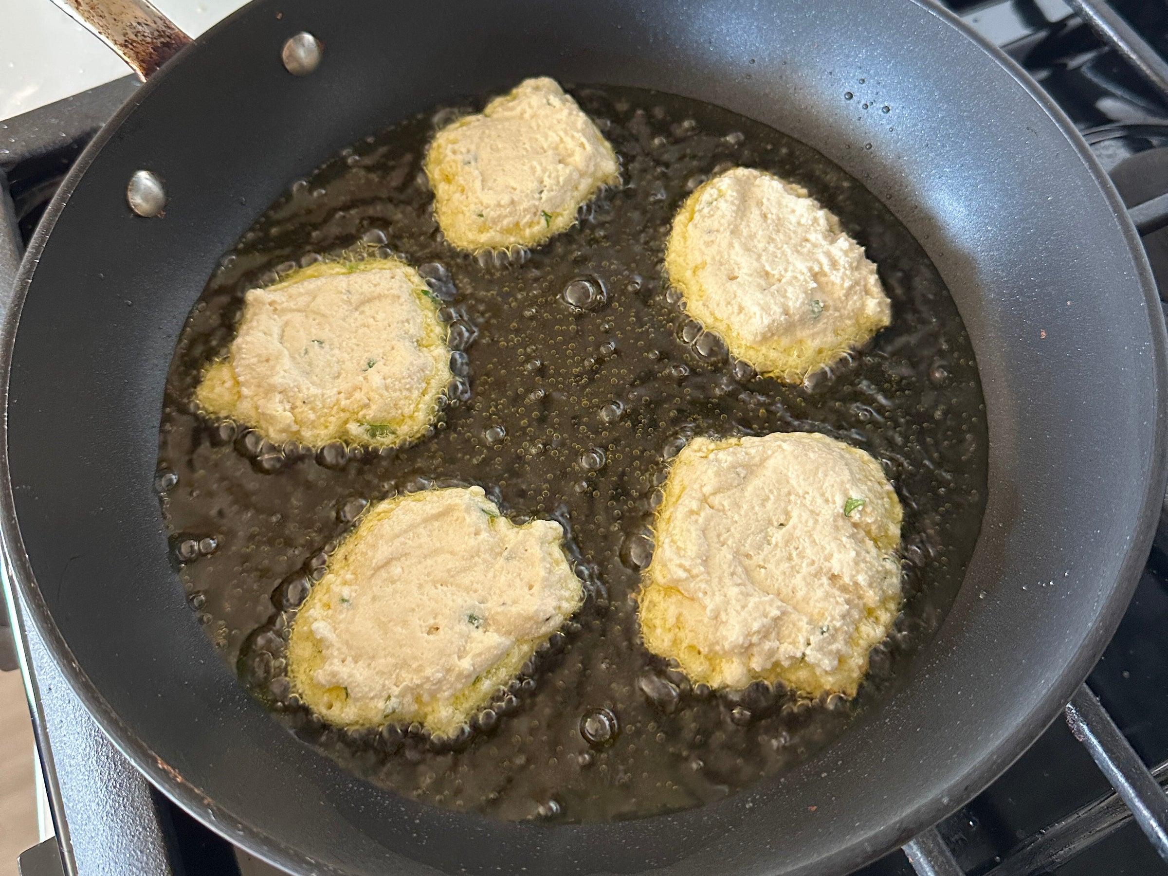 Five patties of batter are frying in hot oil in a black skillet on a stove, with bubbles forming around them as they cook.